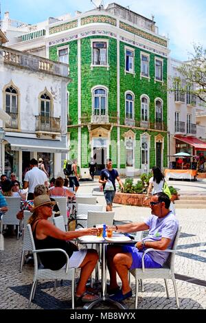 I turisti in un momento di relax a caffetterie in Praça Luis de Camoes con piastrelle verdi tradizionale edificio Portoghese alla parte posteriore, Lagos, Algarve, Portog Foto Stock