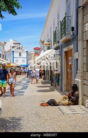 Giocatore seduto sul lastricato della strada dello shopping a suonare la chitarra lungo R 25 de Abril nella città vecchia con turisti di passaggio, Lagos, Algarve, Porto Foto Stock