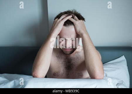 Preoccupato uomo seduto sul letto con la mano sulla fronte. Egli ha problemi di salute mentale e sentire le voci nella sua testa Foto Stock