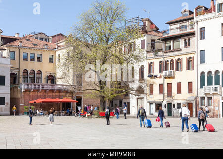 Campo San Polo, San Polo, Venezia, Veneto, Italia in primavera con i turisti tirando le loro valigie attraverso la piazza e la gente del luogo rilassante sotto gli alberi Foto Stock