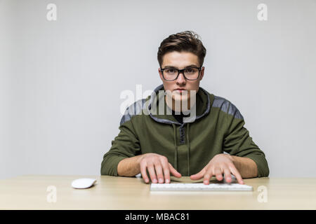 Un giovane hacker con gli occhiali vestito in abiti casual seduti ad una scrivania e lavorare sulla tastiera di un computer nella parte anteriore Foto Stock