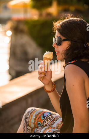 Giovane e attraente donna mangiare gelato al lago durante il tramonto con occhiali da sole Foto Stock