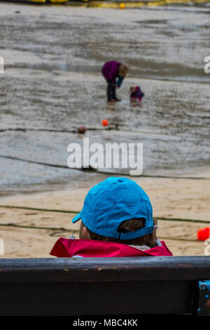Un vecchio o anziano uomo seduto in riva al mare su un banco che si affaccia sul porto di persone a giocare sulla spiaggia. Indossando un blu berretto da baseball. Foto Stock