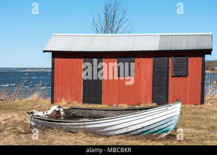 Bianco torbido canotto sul terreno di fronte a un rosso e nero tettoia in legno nel paesaggio costiero. Ubicazione Farjestaden su Oland, Svezia. Foto Stock