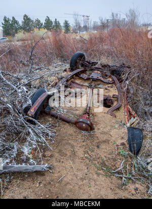 Vecchio arrugginito carro automatica parzialmente sepolti nella sabbia da un vicino torrente. Un contrasto con il nuovo edificio in costruzione la distanza, Colorado. Foto Stock