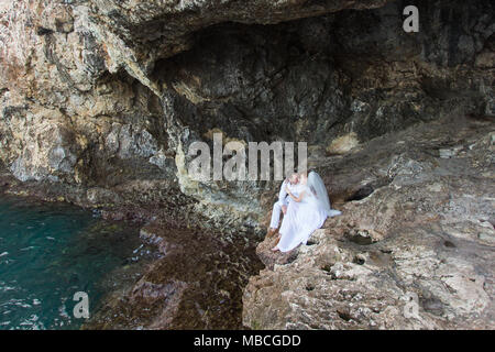 Giovane sposi sposa e lo sposo ride e sorrisi a ciascun altro, felice e gioioso momento. L uomo e la donna nel matrimonio abiti sedersi sulla roccia dello sfondo. Foto Stock