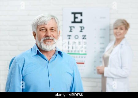 Paziente vecchio di stare di fronte oculista. Guarda soddisfatta. Indossare maglietta blu. cercando di migliorare la salute e la visione. Medico dietro alla ricerca cordiale e expirienced. Foto Stock