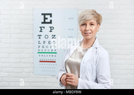 Ritratto di esperto oculista rimanendo davanti a ispezione visiva tabella, appesa sul muro bianco nel laboratorio di analisi mediche Expirienced medico persona aiuta a migliorare la loro salute degli occhi e visione. Foto Stock