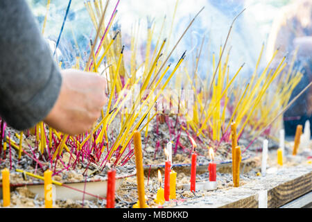 Vista ravvicinata di un maschio di mano di piantare un brucia incenso stick tra monete in sabbia su di un tavolo di legno con le candele accese durante una cerimonia buddista. Foto Stock