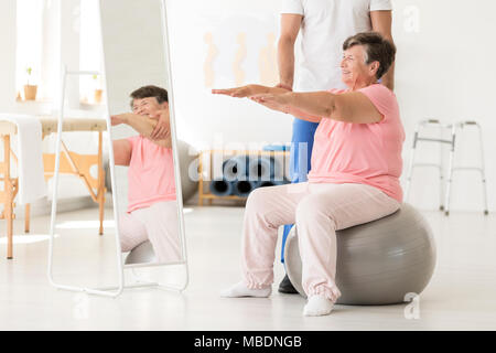 Sorridente senior esercitando con un fisioterapista su una sfera di argento in una palestra bianca Foto Stock