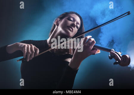 Bella giovane donna a suonare il violino su sfondo blu scuro. La nebbia in background. Studio shot Foto Stock