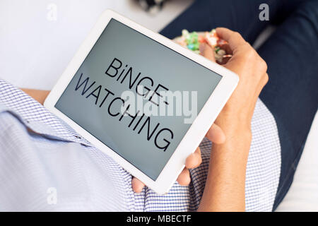 Primo piano di un giovane uomo caucasico comodamente sdraiato su un lettino, mangiare popcorn mentre si sta guardando il suo tablet con il testo binge-watching nella relativa schermata Foto Stock