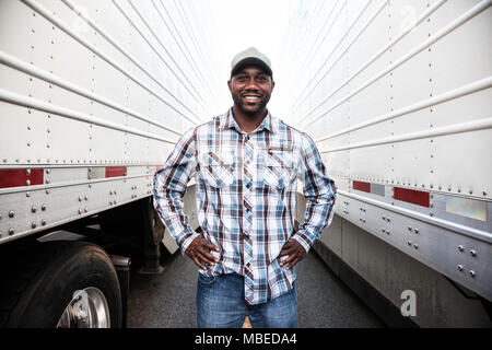 Un camionista che stava fra due grandi rimorchi dei camion in un parco del rimorchio, le mani sui fianchi, sorridente, abiti da lavoro Foto Stock