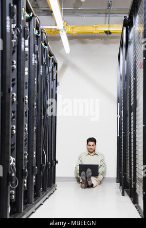 Calcolatore maschio tecnico che lavora su un computer portatile mentre è seduto in un corridoio di server in rack in un computer server farm. Foto Stock