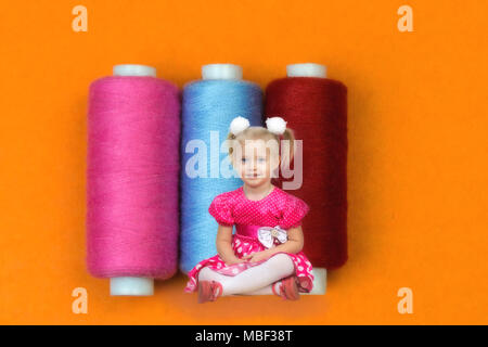 La ragazza in abito rosso si siede attorno a tre grandi bobine con fili di cucitura. Foto Stock