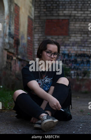 Una ragazza con corti capelli scuri, occhiali, un My Chemical Romance t-shirt e una sigaretta seduto accanto a un vecchio edificio. Foto Stock