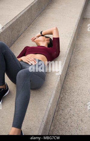 Esaurito sportivo giovane donna sdraiata su scale nel fitness abbigliamento con la sua mano sul suo viso Foto Stock