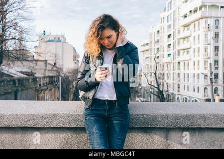 Attraente giovane donna si erge su un ponte e si guarda in uno smart phone tenendolo in mano. Curly redhead girl scrivere messaggi su sfondo di th Foto Stock