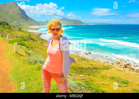 Bionda turista femminile sul popolare attrazione Chapmans Peak Drive nella Penisola del Capo vicino a Cape Town, Sud Africa. Lo stile di vita di viaggiatori donna nel giorno di viaggio su strada. Vacanze estive. Blue sky. Foto Stock