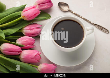 Nero caffè in tazza bianca con tulipani rosa sulla pietra di luce sullo sfondo. Vista superiore Foto Stock