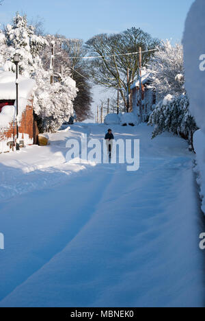 Snowy scena di strada. Villaggio nello Yorkshire. Strada coperta in una profonda copertura di neve su un soleggiato inverni giorno. Persona solitaria a spasso a Natale. Foto Stock