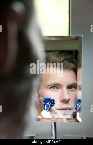 La riflessione di uomo bello con gli occhi verdi la rasatura con il rasoio blu nel piccolo specchio Foto Stock