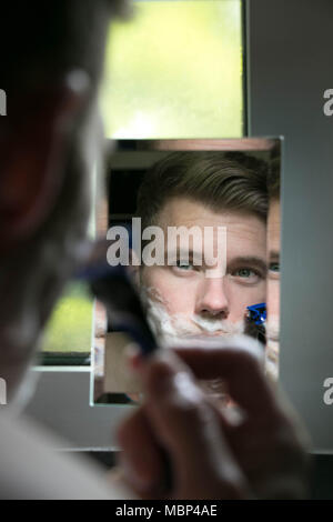 La riflessione di uomo bello con gli occhi verdi la rasatura con il rasoio blu nel piccolo specchio Foto Stock