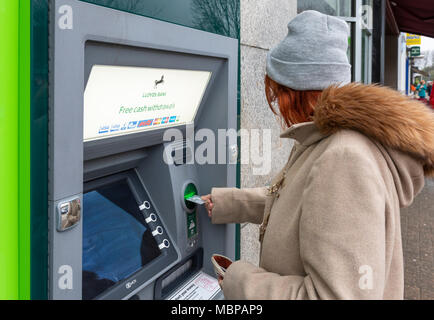 Senior donna prendendo denaro dalla Banca di Lloyds ATM in Inghilterra, Regno Unito. Sportello bancomat, punto cassa, con bancomat, il foro nella parete. Prelievo di denaro. Foto Stock