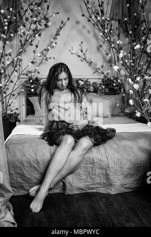 Foto in bianco e nero di cute brunette incinta donna in un abito mantenendo la mano sulla pancia mentre si siede sul letto con fiori sul letto in bedroo Foto Stock