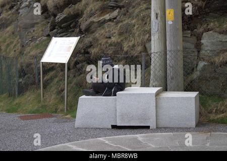 Giovane uomo seduto su lo zoccolo in pietra a Mizen Head attrazione turistica giocando sul suo telefono. Foto Stock