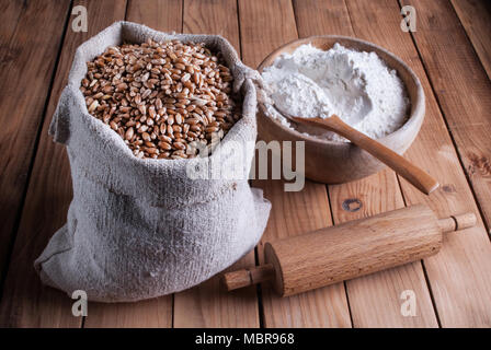 Chicchi di grano in sacchetto di tela e farina bianca nella ciotola di legno e perno di rotolamento sulla scrivania Foto Stock