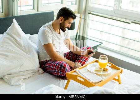 Giovane uomo di mangiare la colazione a letto mentre si utilizza il telefono cellulare Foto Stock