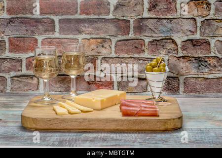 Due bicchieri di vino bianco su un tavolo con il prosciutto, il formaggio e le olive su un sfondo mattone Foto Stock