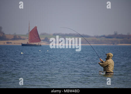 Holnis, Germania - 8 Aprile 2018 - pescatore cintura permanente-profonde nell'acqua fuori dalla spiaggia con la classica barca a vela in background Foto Stock