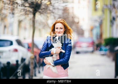 Soggetto uomo e cane. Un rosso giovane con capelli donna caucasica con lentiggini sulla faccia detiene in bianco e nero di shaggy chihuahua cane di razza. La ragazza vestita di bl Foto Stock