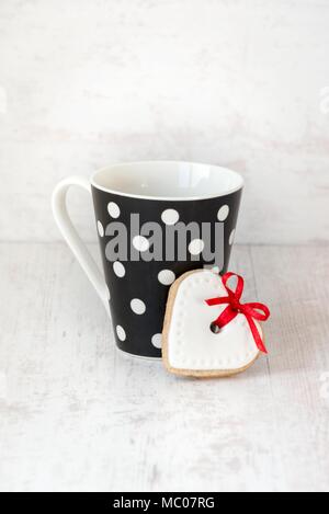 Nero punteggiato di polka tazza di caffè con una a forma di cuore biscotti fatti in casa bianco su uno sfondo di legno. Foto Stock