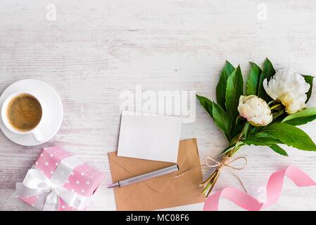 Vista superiore della nota vuota, kraft busta, la tazza di caffè e peonia fiori su legno bianco sfondo rustico.Copia dello spazio. Foto Stock