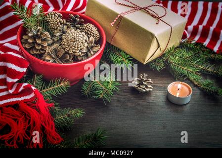 Decorazione di natale - rosso ciotola piena di abeti-coni, confezione regalo avvolti in carta kraft, rami di pino, candela rossa e bianca a strisce inverno sciarpa. Chri Foto Stock