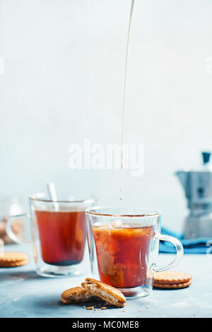 Il caffè del mattino con i cookie. Doppia parete le coppe di vetro con una moka pot su un calcestruzzo leggero sottofondo. Versare il latte nel caffè azione fotografia di cibo Foto Stock