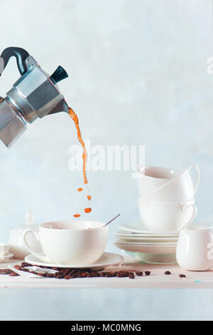 Versare il caffè da una moka pot in una tazza di porcellana. Bianco su bianco ancora in vita con una mensola da cucina, piatti, piattini, bicchieri e spazio di copia Foto Stock