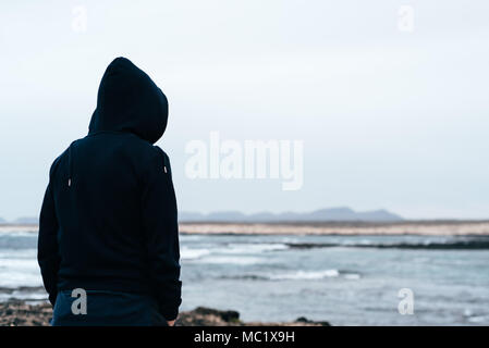 Uomo che indossa hoodie guardando il mare. Vista posteriore del concetto di solitudine Foto Stock