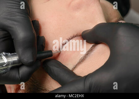 Vista da sopra del professional cosmetologo indossando guanti neri rendendo eyeliner permanente trucco per donna. Client sdraiato sul lettino al salone di bellezza w Foto Stock