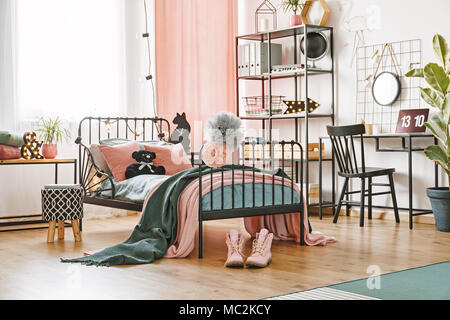 Nero orsacchiotto posto su un letto di metallo con lenzuola e coperte in piedi nella luminosa sala interna con accenti rosa Foto Stock