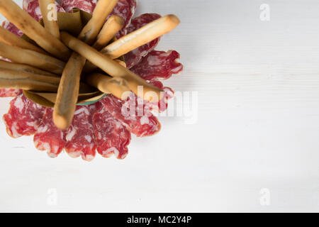 Fette di salame realizzate nel Nord Italia con bastoncini di pane pronto per un antipasto informale su una bianca tavolo in legno, soggetto nell'angolo superiore sinistro Foto Stock