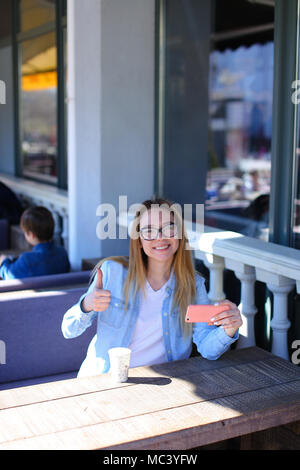 La donna che mostra Thumbs up, seduto alla street cafe con lo smartphone. Foto Stock