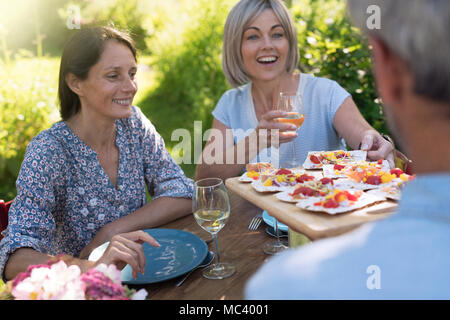 In estate. gruppo di amici in quarantenni si sono riuniti intorno ad un tavolo in giardino per condividere un pasto. Un uomo offre i prosciutti di toast per gli ospiti Foto Stock