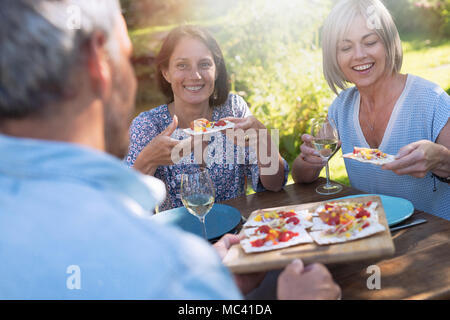 In estate. gruppo di amici in quarantenni si sono riuniti intorno ad un tavolo in giardino per condividere un pasto. Un uomo offre i prosciutti di toast per gli ospiti Foto Stock