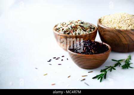 Jasmine riso bianco, marrone, nero e rosso di riso. Assortimento misto di grani in ciotole di legno sul cemento grigio Sfondo, spazio di copia Foto Stock