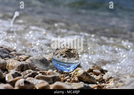 Palla di vetro seduto alla spiaggia rocciosa whit mare onda riflessa in esso/ immagine concettuale delle vacanze estive Foto Stock