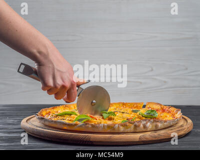 Acciaio pazza taglierina per il taglio di gustoso piatto italiano su supporto in legno isolato. donna di lato con un coltello tagliare la pizza su sfondo bianco di close-up Foto Stock